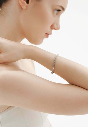 Mujer de piel clara que lleva una pulsera de cadena plateada, apoyando su brazo sobre el hombro y mirando hacia la derecha sobre un fondo blanco.