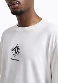 T-shirt bianco in cotone con logo a diamante nero e testo "AX" e "ARMANI EXCHANGE" sottostante. Design a maniche corte e collo rotondo.