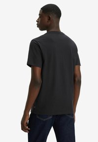 Camiseta de algodón negra con mangas cortas, cuello redondo y una sutil etiqueta de la marca cerca del dobladillo inferior, combinada con unos pantalones vaqueros oscuros.