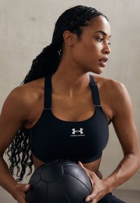 Crni sportski grudnjak s podesivim trakama, s bijelim logotipom Under Armour. Model drži crnu medicinsku loptu koja prikazuje teksturirane detalje.