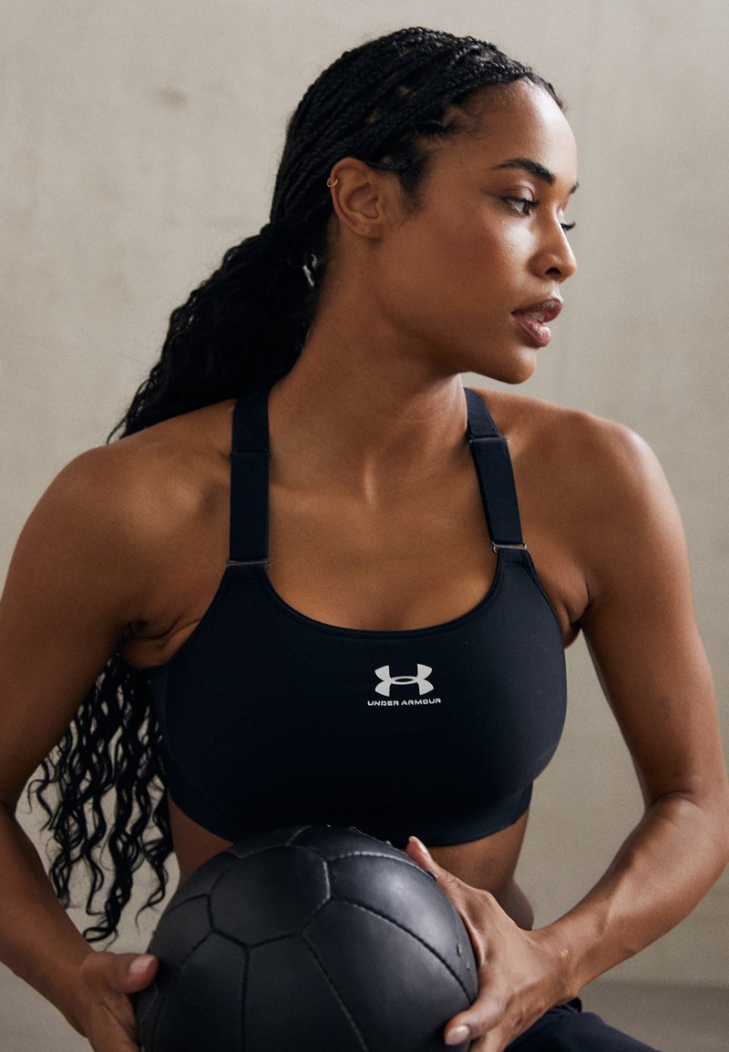 Crni sportski grudnjak s podesivim trakama, s bijelim logotipom Under Armour. Model drži crnu medicinsku loptu koja prikazuje teksturirane detalje.