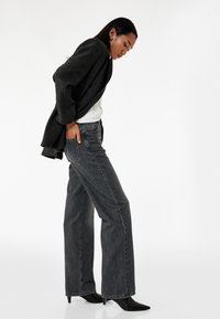 Jeans de mezclilla gris de pierna ancha con cintura alta, combinados con un blazer negro texturizado y tacones negros de punta, sobre un fondo liso.