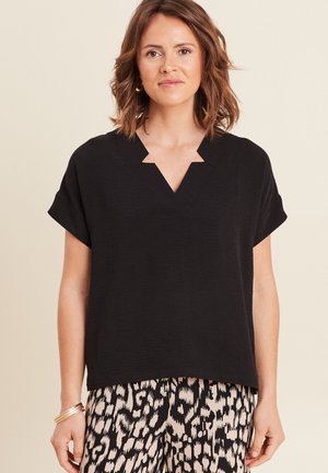 Frau, die ein schwarzes Kurzarm-Oberteil mit V-Ausschnitt und schwarz-beige gemusterte Hose trägt, steht vor einfachem Hintergrund.