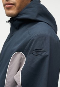 Άνδρας που φοράει σκούρο μπλε μπουφάν με κουκούλα, με γκρι μπαλώματα στο μανίκι και κεντημένο οβάλ λογότυπο στον άνω βραχίονα.