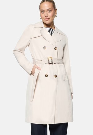 Femme portant un trench-coat beige à double boutonnage avec ceinture, chemise rayée, boucles d'oreilles en or en forme d'anneau, et main dans la poche du manteau.