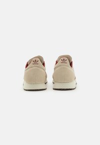 Zapatillas de ante beige con acentos rojos, detalles de malla y suela blanca texturizada. Presenta un logo en el talón y líneas suaves en el diseño.