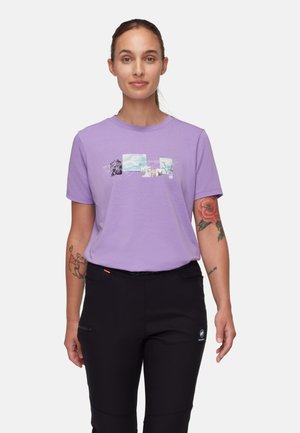 Kobieta z włosami związanymi do tyłu, ubrana w lawendowy t-shirt z nadrukiem i czarne spodnie, stoi na białym tle, z tatuażami na ramionach.