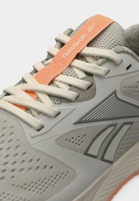 Ανοιχτό γκρι αθλητικό παπούτσι Reebok με λευκά κορδόνια, διάτρητο επάνω μέρος και γλώσσα με ροδακινί λουράκι με το λογότυπο.