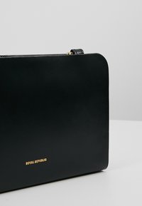 Borsa in pelle nera con una texture liscia, forma rettangolare e logo in rilievo dorato. Presenta un design semplice con una tracolla sottile.