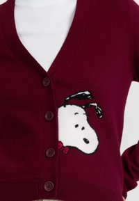 Bordeaux cardigan met een V-hals, voorzien van vijf knopen en een Snoopy-afbeelding op de linkerkant van de borst, gemaakt van zachte breimateriaal.