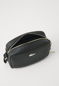 Crna kožna torbica za crossbody s teksturiranom površinom, zlatnim zatvaranjem na munju i Lacoste logotipom u zlatu sprijeda. Pravokutnog oblika.