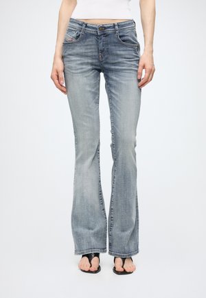 Torso inférieur d'une personne portant un jean bootcut bleu clair et des sandales noires à bouts ouverts, debout devant un fond clair uni.