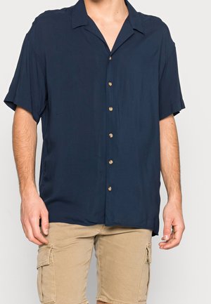 Homme portant une chemise bleu marine à manches courtes boutonnée et un short cargo beige sur un fond uni.