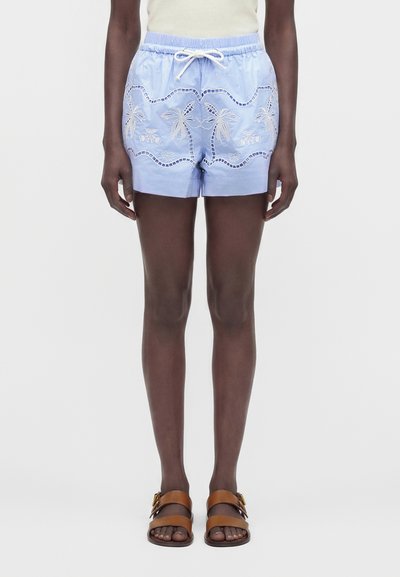 Personne portant un short bleu clair avec des palmiers et des vagues brodés, associé à des sandales marron à double bride et un haut blanc.