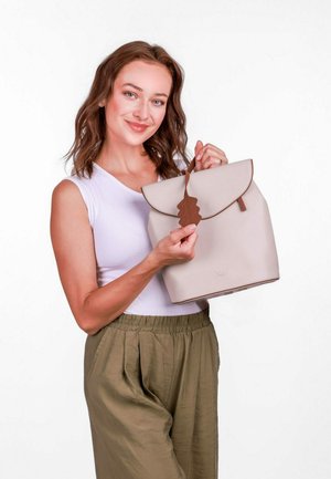 béžový batoh s přehnutým klopovým víkem, hnědou akcentující částí ve tvaru listu a hladkým povrchem, drží osoba oblečená v bílém topu a olivových kalhotách.