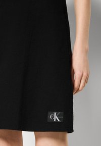 Μαύρο φόρεμα από υφασμάτινη υφαντική ύλη, με ετικέτα λογότυπου στο άκρο που αναγράφει "Calvin Klein Jeans" σε άσπρο και μαύρο χρώμα. Απλή, ευθεία γραμμή.