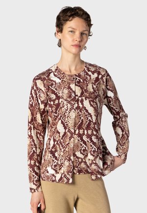 Kvinde med kort hår iført langærmet, knappet skjorte med brun og beige slangeprint, kombineret med beige bukser mod en ensfarvet baggrund.