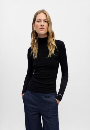 Jeune femme aux longs cheveux blonds portant un pull à col roulé noir et un pantalon bleu foncé, debout devant un fond gris clair uni.