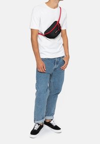 Black and red nylon crossbody bag worn over a white t-shirt, paired with blue denim jeans and black sneakers with white laces.