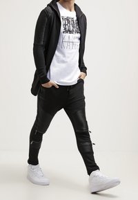 Svart zip-hoodie med texturerade axelpaneler över en vit grafisk t-shirt, kombinerad med svarta joggers med dragkedjefickor och vita sneakers.