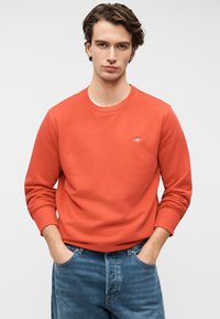 Κόκκινο φούτερ με στρογγυλή λαιμόκοψη και ριμπ μανσέτες, με μικρό λογότυπο στο στήθος. Συνδυάζεται με μπλε τζιν παντελόνι.