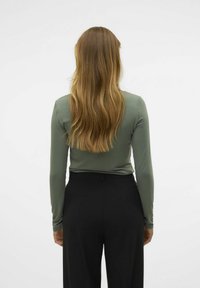 Top ajustado de manga larga en un tejido verde apagado, combinado con pantalones negros de tiro alto, visto desde atrás. Textura suave, diseño casual.