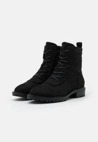 Bottes chevilles en suède noir à lacets, avec des semelles en caoutchouc épaisses et des talons bas, placées côte à côte sur un fond blanc.