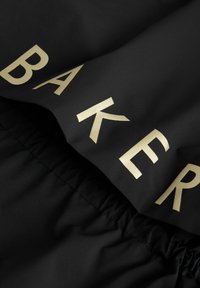 Sort stof med en glat tekstur, der har store, fede guld bogstaver, der staver "BAKER." Inkluderer indsamlet kantdetalje langs kanten.