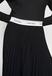 Jupe noire plissée avec top noir côtelé, dotée d'une ceinture blanche imprimée du logo "Calvin Klein" répété.