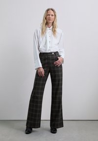 Camisa blanca de manga larga con cuello de pajarita, combinada con pantalones de cuadros de tiro alto y pierna ancha en tonos oscuros y un acento de botón plano.