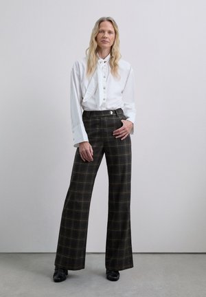 Camisa blanca de manga larga con cuello de pajarita, combinada con pantalones de cuadros de tiro alto y pierna ancha en tonos oscuros y un acento de botón plano.