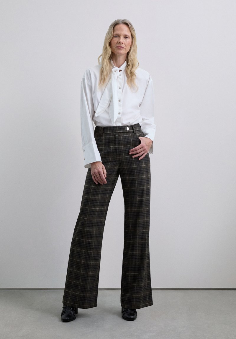 Camisa blanca de manga larga con cuello de pajarita, combinada con pantalones de cuadros de tiro alto y pierna ancha en tonos oscuros y un acento de botón plano.