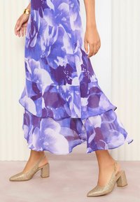 Violetti kerroksellinen kukkamekko, jossa on röyhelöitä, yhdistettynä kimaltaviin kultaisiin slingback-korkokenkiin, joissa on lohkarekorko ja kärki.