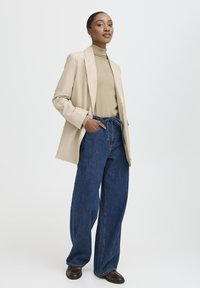 Donna in piedi che indossa un blazer beige e un dolcevita con jeans a gamba larga blu e scarpe marroni scure, con una mano in tasca su uno sfondo neutro.