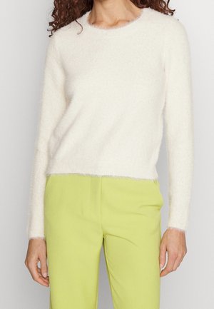 Femme portant un pull blanc duveteux à manches longues et un pantalon vert clair taille haute, visible du cou aux hanches.