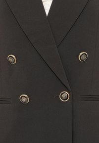 Svart dobbeltrad blazer laget av glatt stoff, med gullknapper med et skjolddesign og to vertikale lommer.