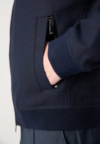 Kéz egy sötétkék dzseki cipzáras zsebében, bordás mandzsettával és derékkal, kék kockás nadrág felett.