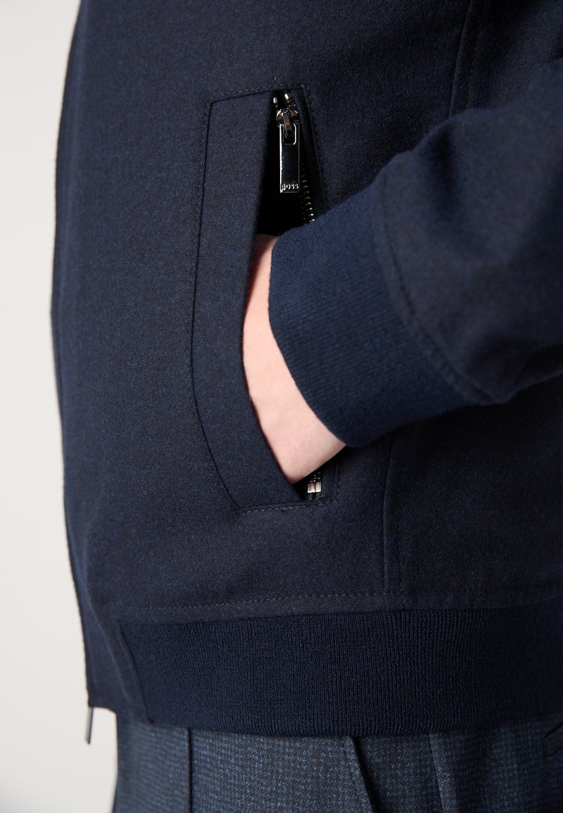 Ręka w kieszeni zamkniętej na zamek granatowej kurtki z prążkowanymi mankietami i pasem, noszonej na niebieskich spodniach w kratę.