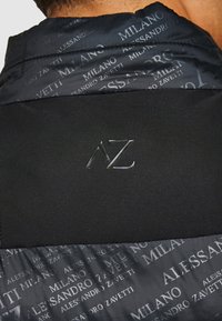 Giacca a vento nera con un tessuto testurizzato che presenta un motivo a logo ripetuto. Un logo "AZ" in rilievo è posizionato sul pannello nero uniforme.