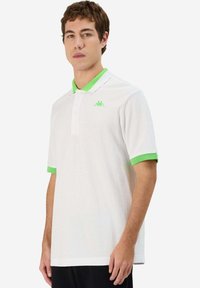 Polo shirt bianco in cotone, con colletto e polsini verdi, dotato di un piccolo logo verde sulla parte sinistra del petto.