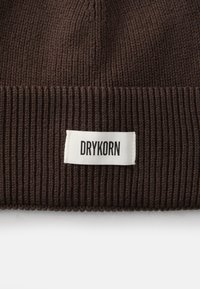 Gorro de malha canelada castanho com uma etiqueta branca que apresenta o texto "DRYKORN". A textura é suave e a forma é arredondada na parte superior.