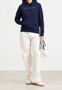Sudadera con capucha navy con el logo "TOMMY JEANS" en blanco, pantalones de pierna ancha crema, zapatillas blancas y un pequeño bolso blanco con acentos navy.