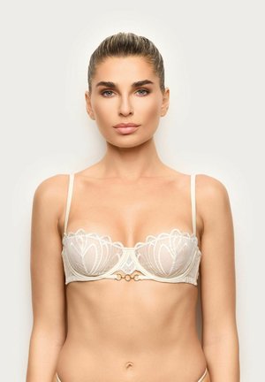 Woman with pulled-back hair wearing a sheer white lace bra with thin straps, facing forward against a plain light background.