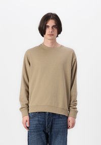 Beige pulóver hosszú ujjakkal, kerek kivágással és bordázott mandzsettákkal. Sötétkék farmernadrággal párosítva, minimalista dizájnnal.
