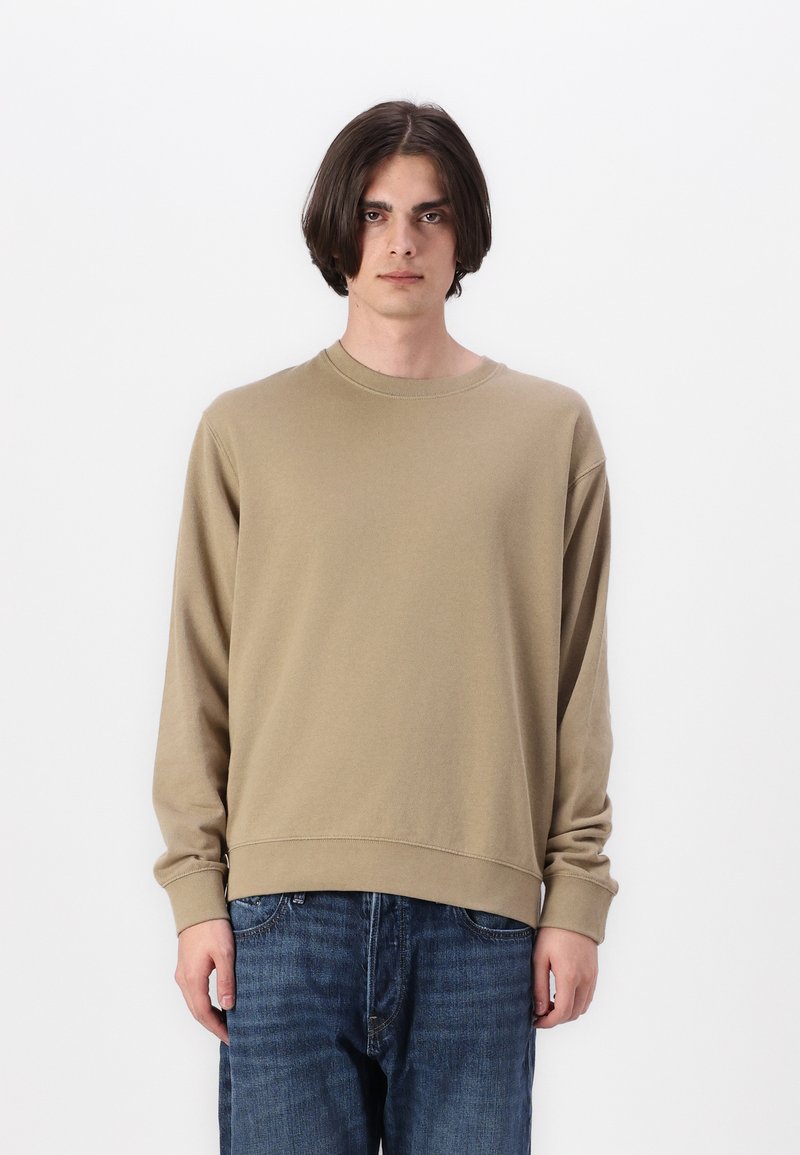 Beige pulóver hosszú ujjakkal, kerek kivágással és bordázott mandzsettákkal. Sötétkék farmernadrággal párosítva, minimalista dizájnnal.