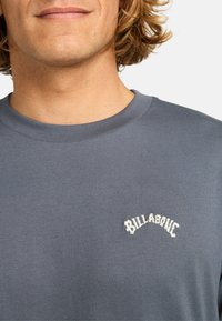 Šedá bavlněná mikina s kulatým výstřihem, měkkou strukturou a bílým vyšitým logem "Billabong" na levé části hrudníku.