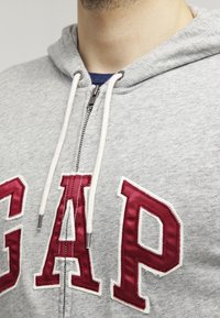 Primer plano de una persona que lleva una sudadera gris de GAP con cordones blancos y letras rojas cosidas en la parte delantera.