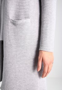 Grå ribbad stickad kappa med långa ärmar och en framficka. Texturen är mjuk, och designen har vertikala vågor genom hela plagget.