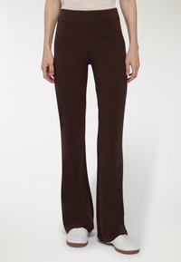 Osoba w ciemno-brązowych prostych spodniach i białych trampkach, stojąca na tle gładkiej białej ściany.