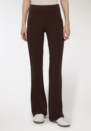 Persona che indossa pantaloni dritti marrone scuro e sneakers bianche, in piedi di fronte a uno sfondo bianco semplice.
